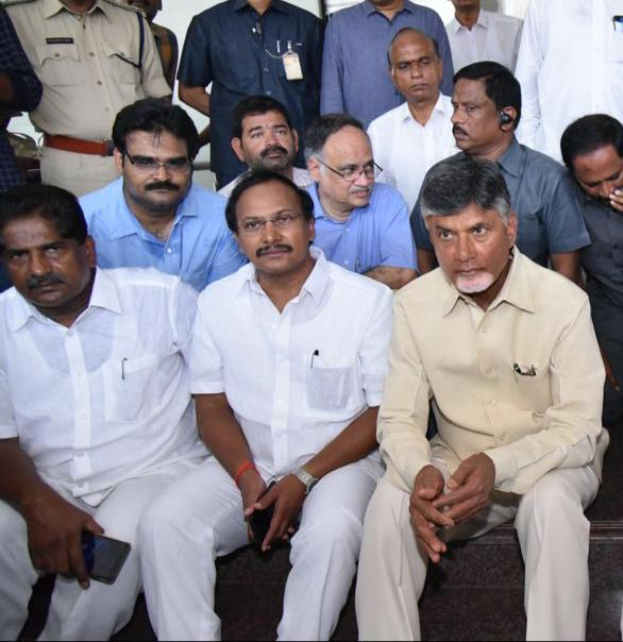 Andhra Pradesh Chief Minister Chandrababu Naidu und seine Kabinettsmitglieder treffen sich zu einer Besprechung in Hyderabad, wobei mehrere Personen auf der Treppe im Vordergrund sitzen und andere im Hintergrund stehen, während ein Text am unteren Rand darauf hinweist, dass die Besprechung an einem öffentlichen Ort stattfindet.
