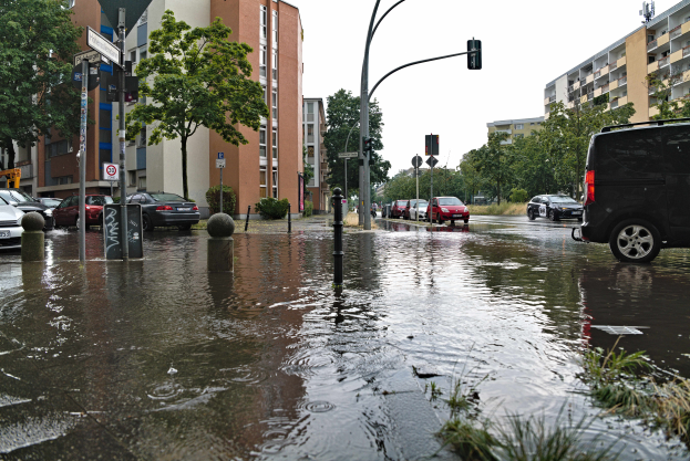 Eine überflutete Straße in Berlin, Deutschland, mit geparkten Autos, Gras, Pfählen, Verkehrsampeln, Schildern, Bäumen, Gebäuden mit Fenstern, unter einem sichtbaren Himmel.