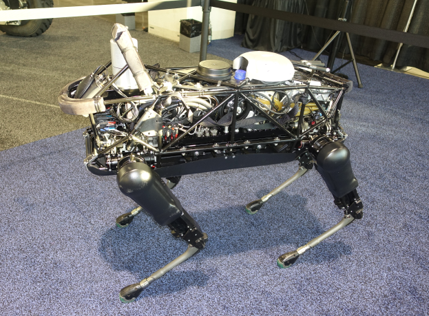 Ein Roboter mit ausgestreckten Gliedmäßen und geneigtem Kopf sitzt auf einem blauen Teppichboden, mit einer Maschine in der Mitte und einem Fahrzeug auf der linken Seite, umgeben von Ständen, Vorhängen und anderen Gegenständen im Hintergrund.