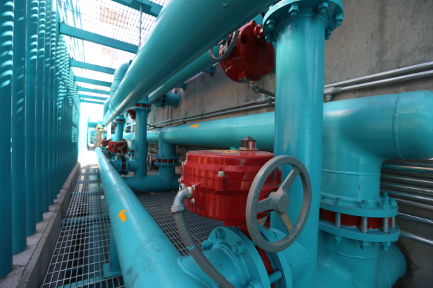 Eine große Gruppe von blauen Rohren und Ventilen in einem Gebäude, mit einem prominenten roten Ventil in der Mitte, Metallstangen links und einer Wand im Hintergrund, wahrscheinlich zeigt das Bild den Bau einer Gasleitung.