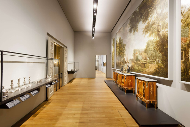 Eine Museumsgalerie mit verschiedenen Kunstwerken, darunter Wandgemälde, Gegenstände auf Tischen, eine Glasvitrine, Informationsschilder und Deckenbeleuchtung.