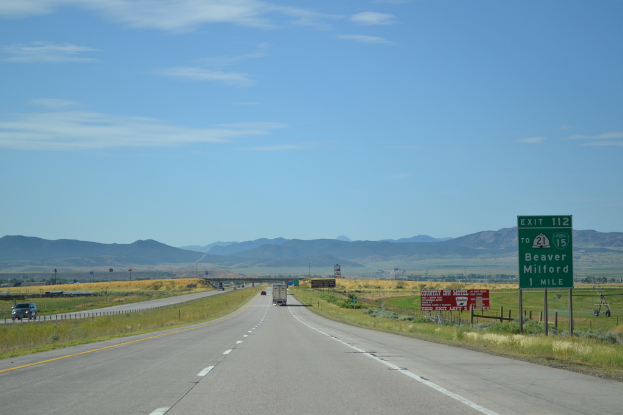 Eine Landschaft mit einem bewölkten Himmel, Hügeln, einer Straße mit Kraftfahrzeugen, Barrieren, Schildern, Informationsanzeigen, landwirtschaftlichen Betrieben und Grasflächen.