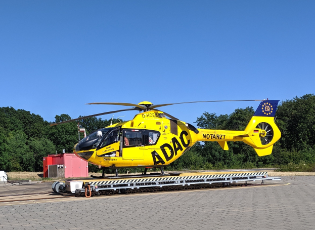 Gelber Hubschrauber auf einem Flughafenvorfeld mit umliegenden Bäumen und einem klaren blauen Himmel, wahrscheinlich für den Luftrettungsdienst in Deutschland verwendet.
