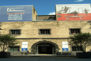 Außenansicht des Deutschen Sport & Olympiamuseums in Berlin mit einem Gebäude mit Fenstern und einer Tür, umgeben von Bäumen und Bannern unter einem bewölkten Himmel.
