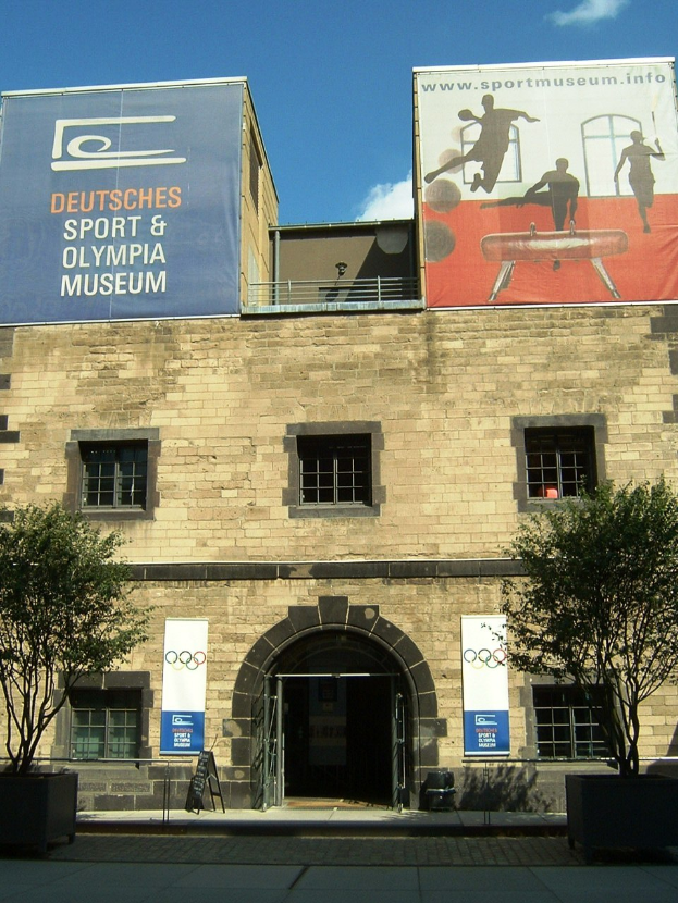 Außenansicht des Deutschen Sport & Olympiamuseums in Berlin mit einem Gebäude mit Fenstern und einer Tür, umgeben von Bäumen und Bannern unter einem bewölkten Himmel.