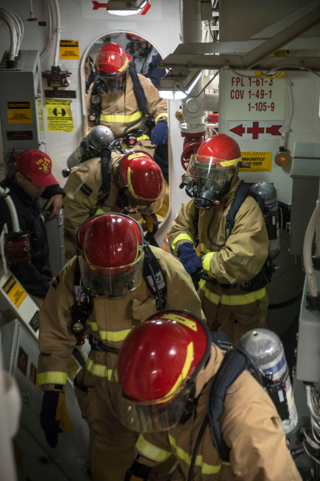 Eine Gruppe von Feuerwehrleuten in Schutzausrüstung, einschließlich Helmen, Handschuhen und Sauerstoffflaschen, arbeitet an einem Boot mit Rohren und anderen Gegenständen im Hintergrund und Plakaten mit Text an der Wand.