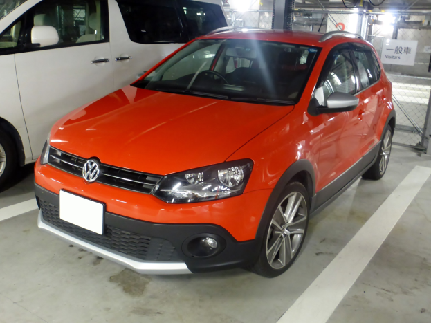 Orange Volkswagen Polo Cross 1.6 TDI Highline DSG neben einem weißen Transporter in einer Tiefgarage geparkt, mit Metallzaun, Hinweisschild, Deckenleuchten und Dach im Hintergrund.