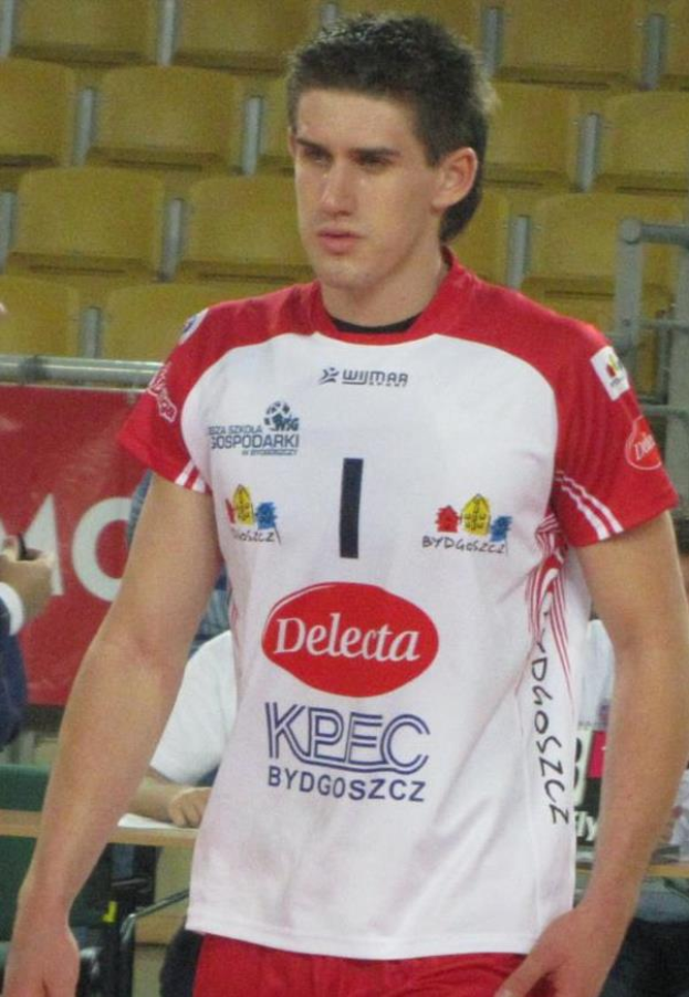 Ein Mann in rot-weißer Handballdress steht auf einem Feld, umgeben von Zuschauern, mit einem roten Banner im Hintergrund, das 'Krasnoyarsk vs Bydgoszcz' zeigt, entschlossen.