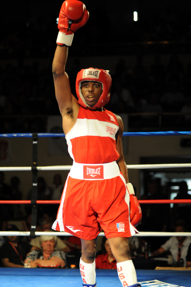 Ein Mann im Boxoutfit, einschließlich Helm und Handschuhen, steht in einem Boxring mit Zuschauern im Hintergrund.
