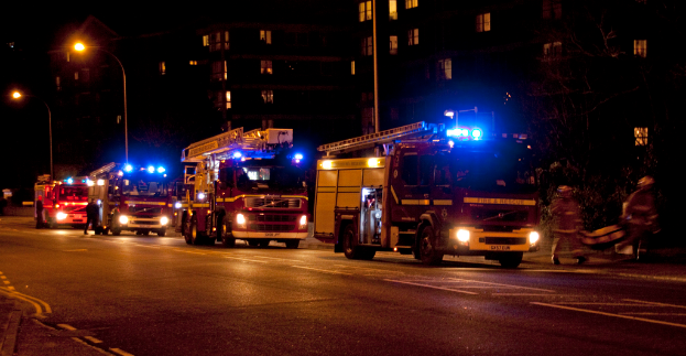 Feuerlöschwagen sind nachts auf einer Straße unterwegs, mit Gebäuden und Laternenmasten im Hintergrund und zwei Personen auf der rechten Seite.