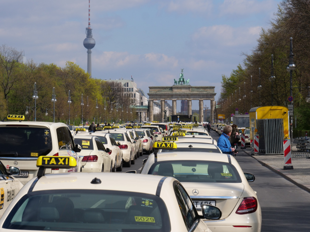 Eine belebte Straße in Berlin mit vielen geparkten Taxis, Fußgängern auf dem Gehweg, Laternen, Bäumen, Gebäuden und einem fernen Bogen mit Statuen und einem Turm unter einem bewölkten Himmel.