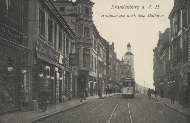 Ein altes Schwarz-Weiß-Foto einer Stadtstraße in Brandenburg, Deutschland, mit einer Straßenbahn auf Schienen, Menschen auf dem Gehweg, Gebäuden mit Fenstern, Schildern und Straßenlaternen unter einem bewölkten Himmel.
