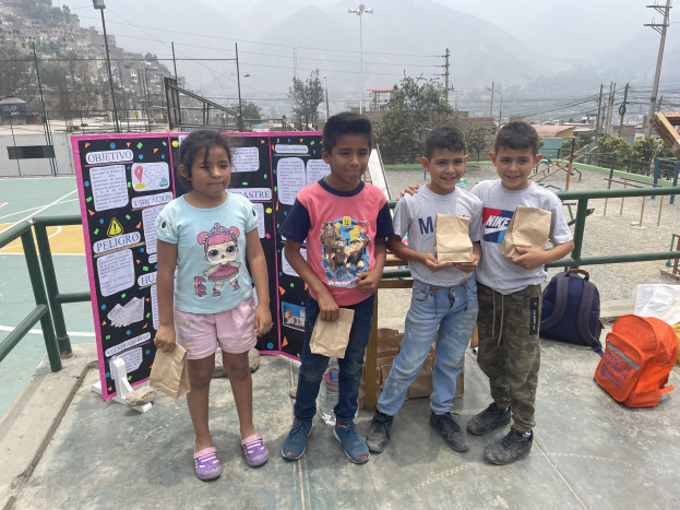 Gruppe von Kindern auf einem Basketballfeld mit Papiertüten in den Händen, Tüten und Gegenstände rechts und eine Tafel mit Text links, vor einem Hintergrund aus Zäunen, Pfählen, Bäumen, Gebäuden, Hügeln und Himmel