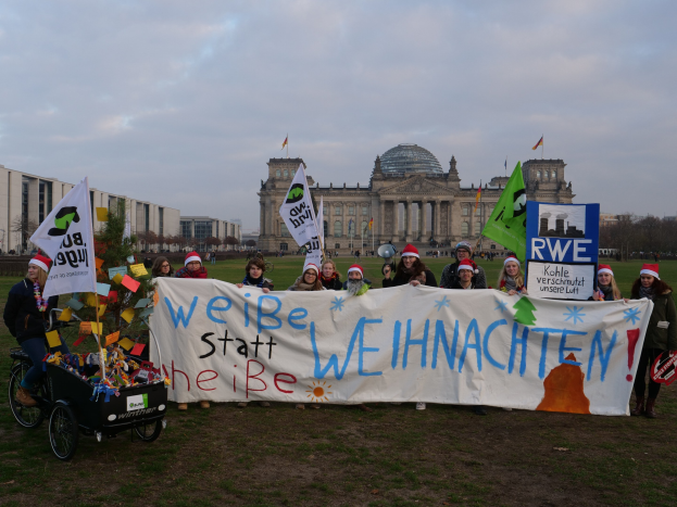 Gruppe von Menschen in Mützen, die ein Banner vor dem Reichstag halten, mit einer Person in einem Kinderwagen, Rasen, Bäumen, Gebäuden, Fahnen und bewölktem Himmel.