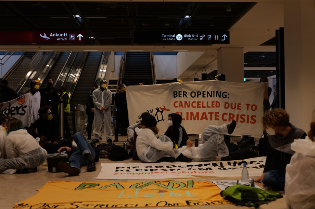 Gruppe von Menschen, die auf dem Boden eines Flughafens sitzen und ein 'Berlin wegen Klimakrise gestrichen' Schild halten, mit verstreuten Gegenständen und Rolltreppen im Hintergrund.