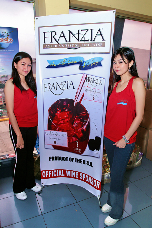 Zwei Frauen neben einem Plakat mit der Aufschrift 'Franzia Americas best selling wine', im Hintergrund ein Glasfenster.