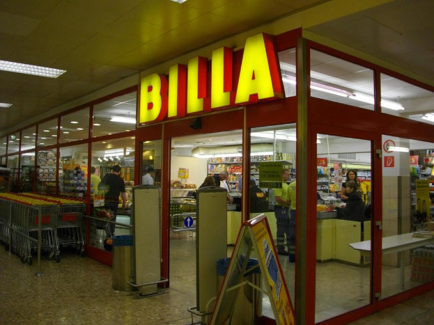 Ein Markt mit einem Eingang in der Mitte, links aneinandergereihte Einkaufswagen und einige Menschen, die im Inneren Produkte anschauen, mit Mülltonnen neben dem Eingang und Lampen oben.