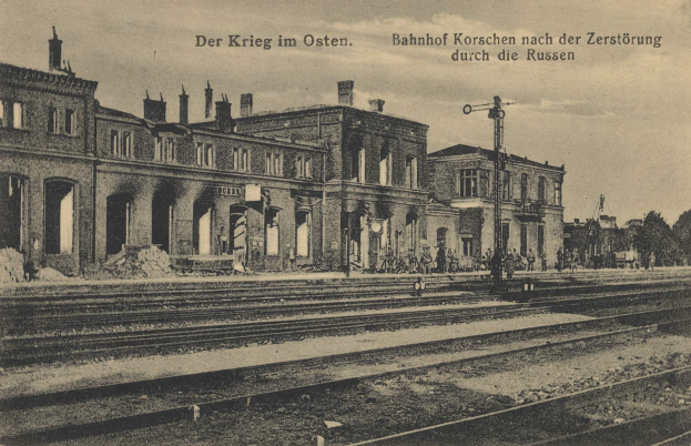 Schwarzes und weißes Foto der Bahnstation Korschen in Ostern, Deutschland, mit Gleisen, Bahnhofsgebäuden, Menschen, Bäumen und Himmel.