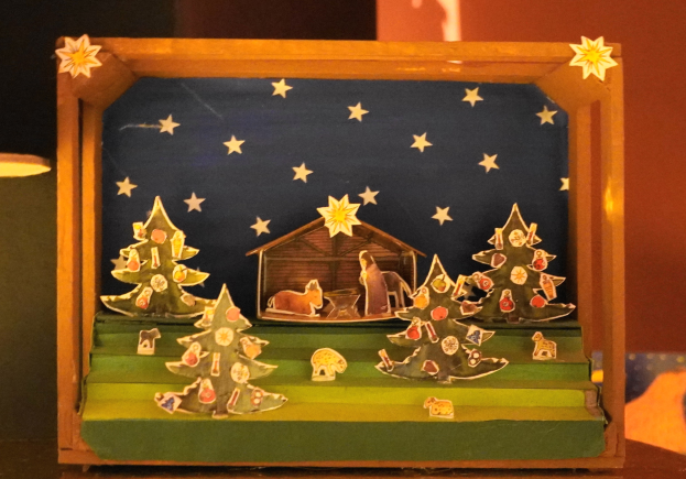 Eine hölzerne Krippe in einer Schachtel auf einem Tisch mit einer Lampe links und einer Wand im Hintergrund, geschmückt mit Weihnachtsbäumen, Tieren und einem Haus.