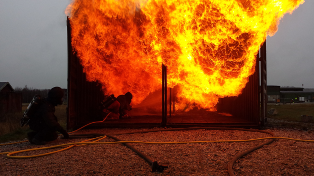 Feuerwehrleute löschen ein Gebäude Feuer mit einer Schlauch auf dem Boden, Schuppen und Bäume sind im Hintergrund unter einem klaren blauen Himmel sichtbar.