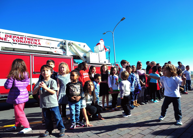 Kinder vor einem Feuerlöschfahrzeug, das für den Weihnachtsumzug der Yokosuka-Feuerwehr beschriftet ist, auf einer Straße mit Laternenpfählen, Bäumen und einem klaren blauen Himmel.