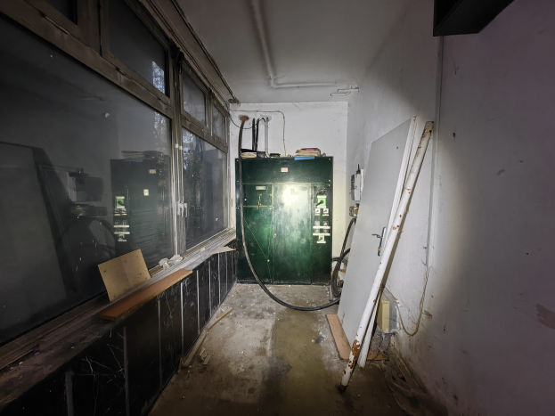 Verlassene Tankstelle mit grüner Tür, Glasfenstern links, Wand rechts, zentraler Maschine und Rohren an den Wänden.