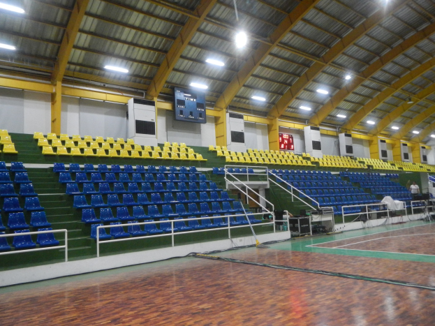 Großer Hallensportplatz mit Holzboden, blauen und gelben Sitzplätzen, Geländern, Treppen mit Geländern, wandmontierten Bildschirmen, Deckenleuchten und einigen Anwesenden.