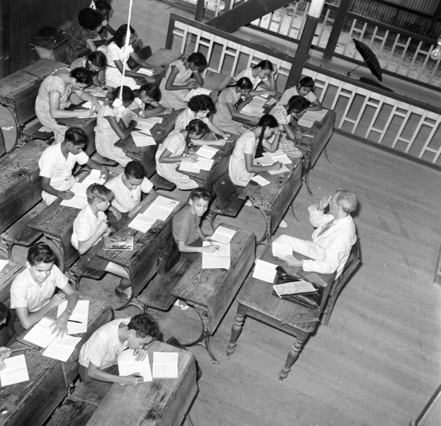 Schwarzes und weißes Foto einer Klasse mit Kindern an Tischen, Büchern und Gegenständen auf den Tischen und einem Geländer und einem Schirm im Hintergrund.