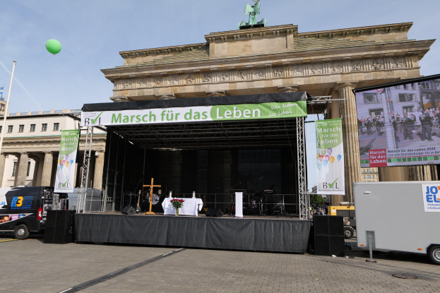 Bühne vor dem Brandenburger Tor mit einem Tisch, der eine Vase, ein Kreuzsymbol, Banner, Lautsprecher und andere Gegenstände enthält, Fahrzeuge auf der Straße, Gebäude mit Fenstern, eine Statue, ein Fahnenmast und ein bewölkter Himmel.