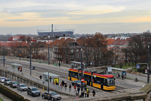 Eine gelbe und schwarze Straßenbahn fährt neben hohen Gebäuden auf einer belebten Stadtstraße mit Autos, Fußgängern, Laternen, Schildern, Bäumen und Gras, unter einem bewölkten Himmel.