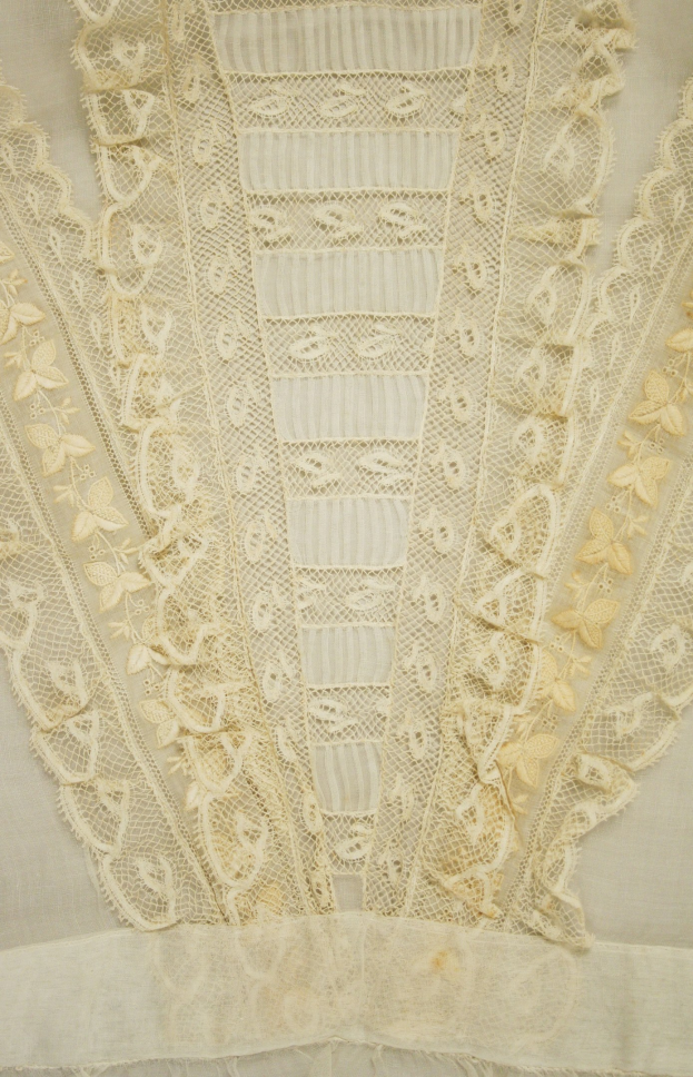 Nahaufnahme eines weißen Kleides mit zarter, detailreicher Spitzenverzierung, die verschiedene Formen und Muster auf einem leichten, luftigen Stoff zeigt.