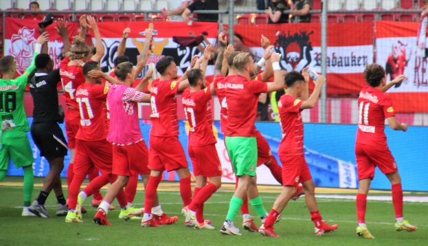 Eine Gruppe von Fußballspielern, die ein Tor feiern, einige halten Flaschen und andere klatschen, mit einem Zaun, Bannern, Metallstangen, Stühlen und einer Decke mit Deckenleuchten im Hintergrund.