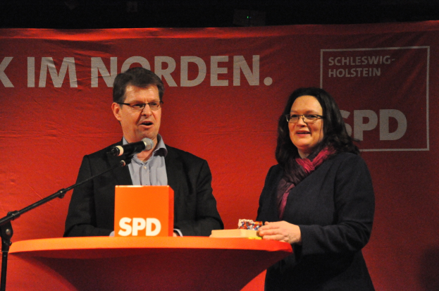 Zwei Personen, die auf einer Bühne vor einem Tisch mit Gegenständen und einem Mikrofon stehen und im Hintergrund ein Banner sehen.