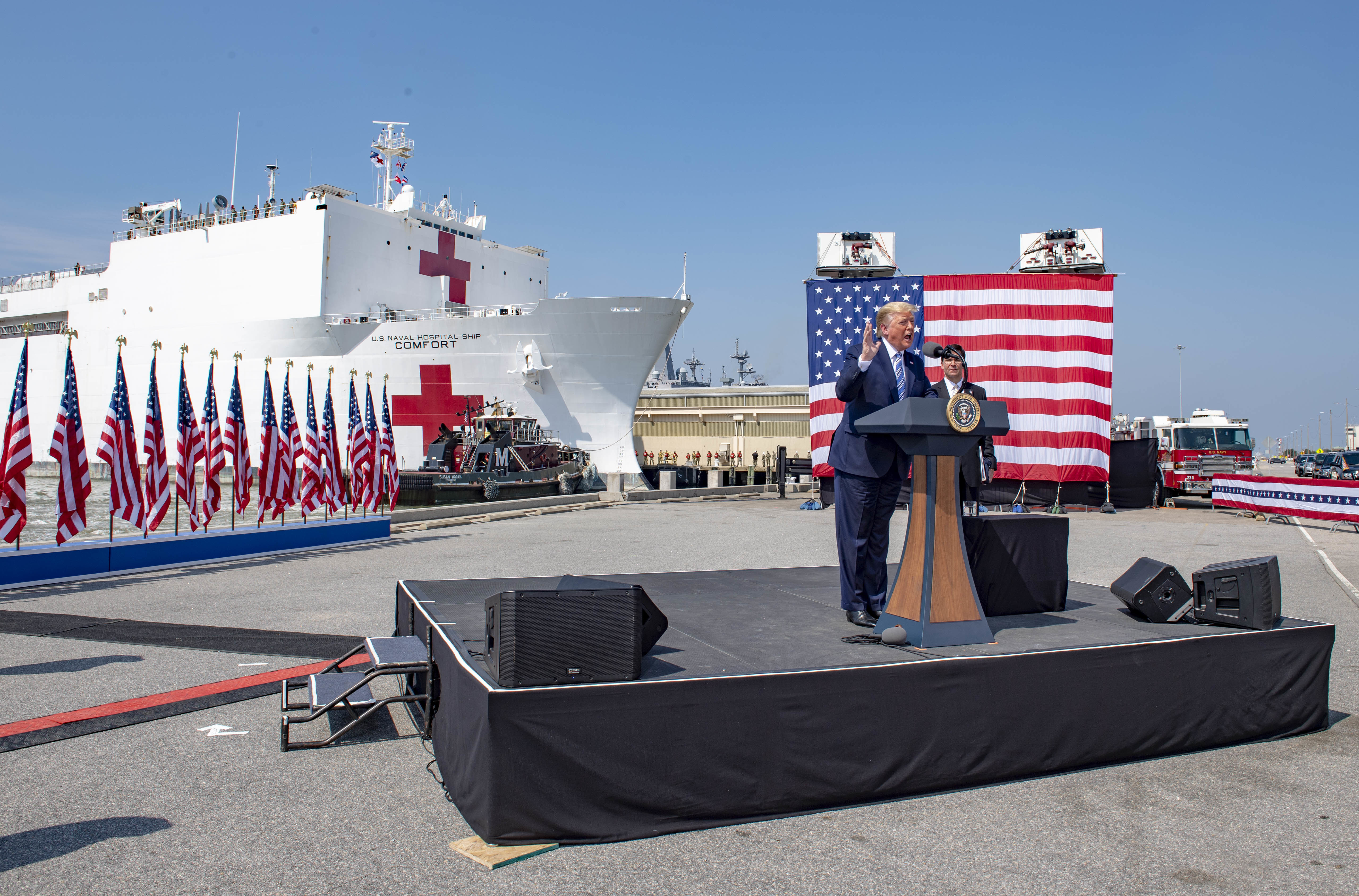 Präsident Donald Trump spricht an einem Rednerpult vor einem großen Schiff, umgeben von Fahnen, Fahrzeugen und anderen Gegenständen, trägt ein Sakko, Krawatte und Hemd und hält ein Mikrofon, mit dem Himmel im Hintergrund.